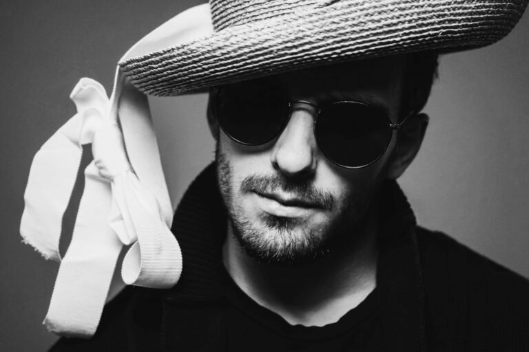 Man with beard wears sunglasses and straw hat with white ribbon in black and white.